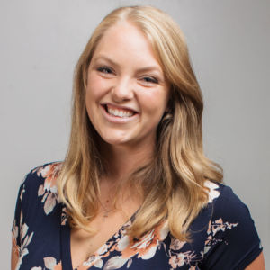 Katie Bertsch smiling headshot wearing a navy blue floral pattern shirt in front of a grey background