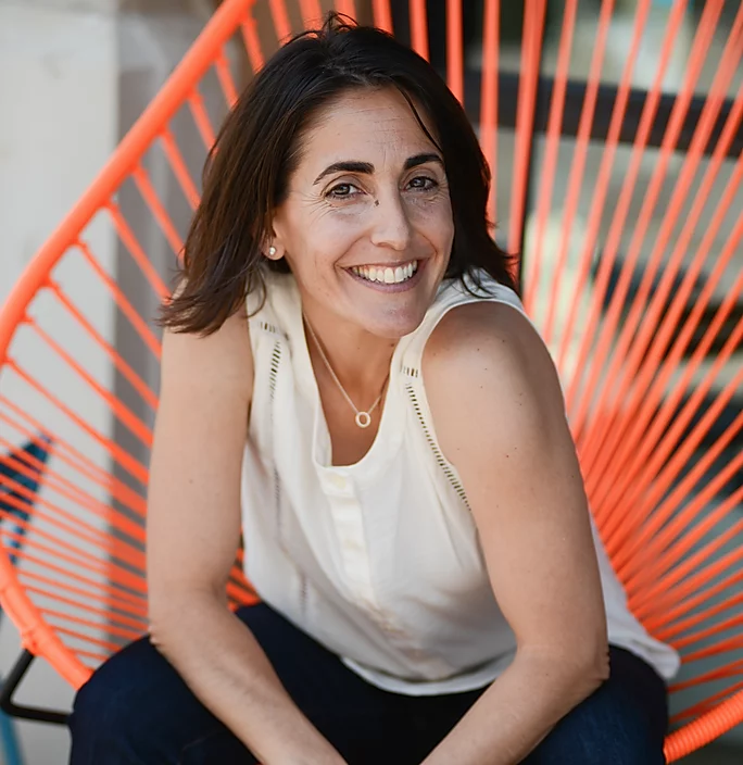 Woman wearing a cream colored tank top and jeans sits on a bright orange chair outside