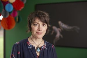 Woman wearing a floral blouse and necklace in front of a green wall with a large canvas photo and hanging paper lanterns in the background