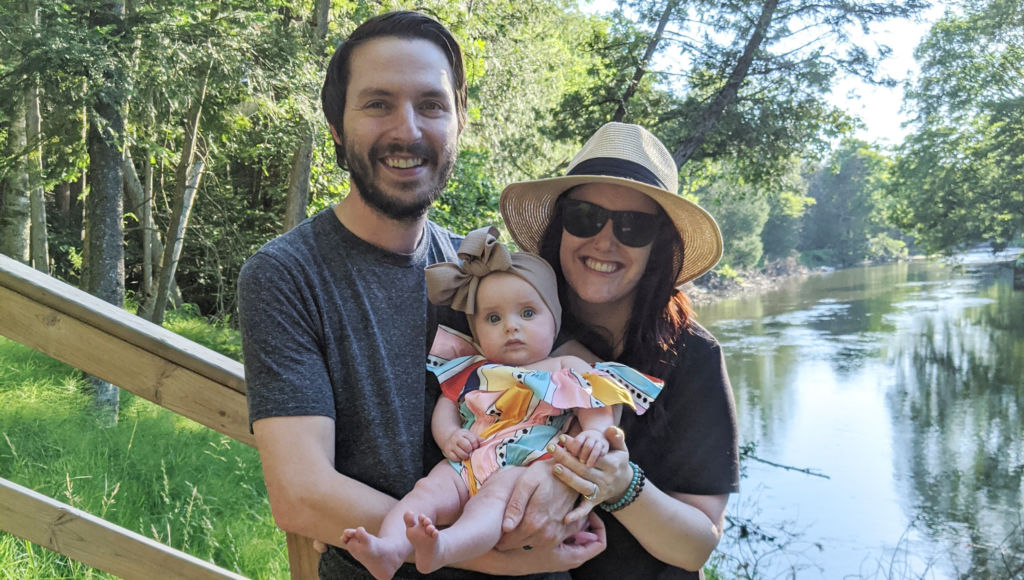 Audra Geyer family photo with husband and infant daughter standing outside on stairs in front of a river and wooded area