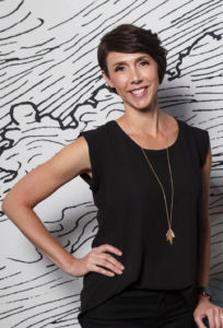 Alyssa Veneklase wearing a black tank top, black pants, and long necklace with hand on her hip in front of a black and white striped wall