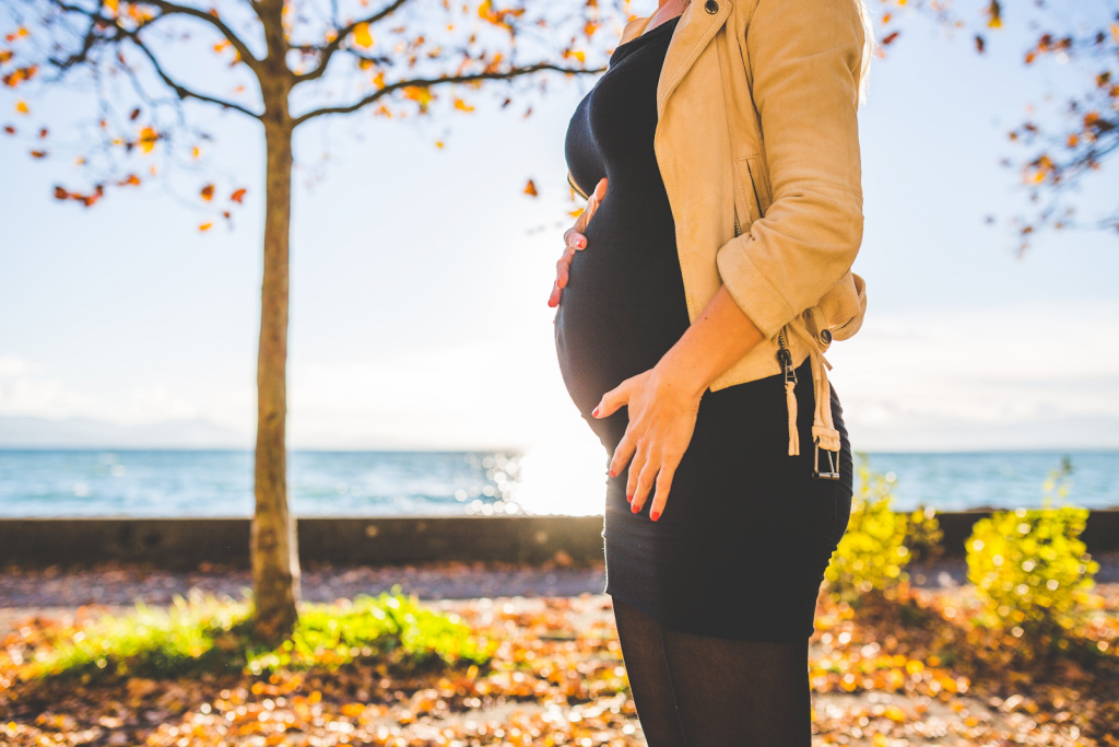 Pregnant woman wearing black dress and mustard yellow jacket standing in front of a body of water, autumn tree, and holding her pregnant belly