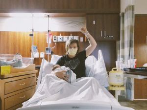 New mom wearing a mask in a hospital bed holds her fist up in victory while holding her newborn