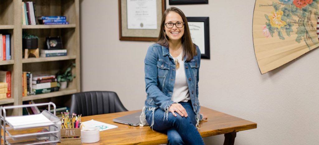 Woman wearing jeans, a white tshirt, and a jean jacket sits on top of a desk in her office