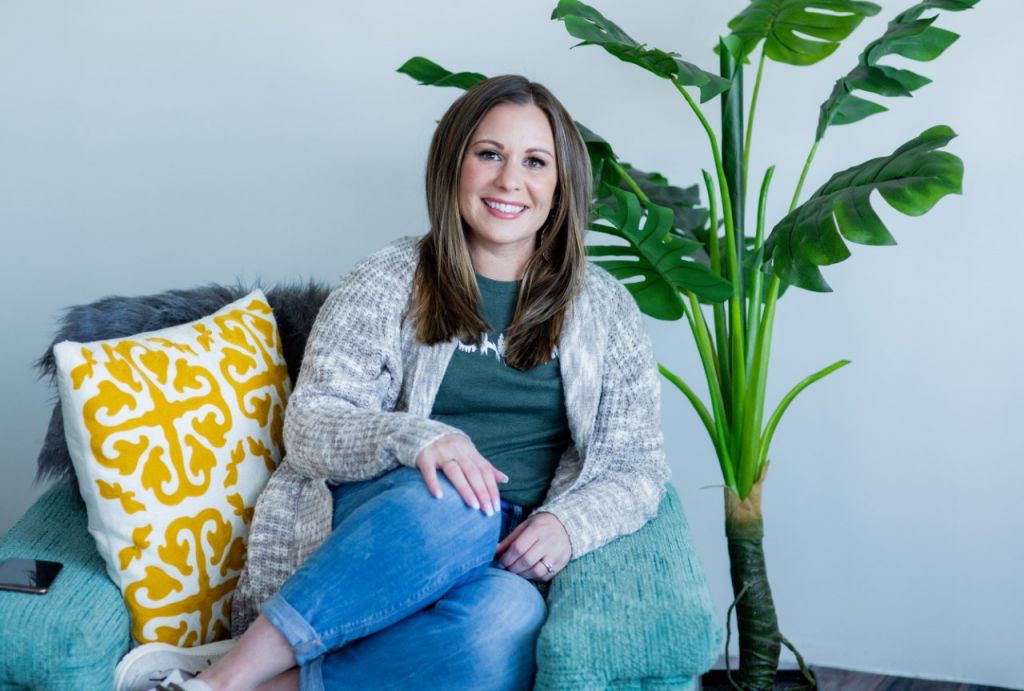 Woman wearing jeans, a teal graphic t, and grey sweater sits in a teal chair with a pillow and blanket beside a house plant