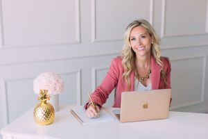 Elizabeth wearing a white blouse with a pink blazer sitting at a white table with a lap top, golden pineapple, flowers, and a pen and paper