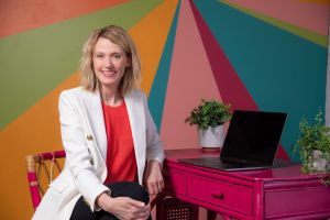 Kristin Revere of Gold Coast Doulas sits at a pink desk with plants and a laptop in front of a colorful wall wearing a coral blouse and white blazer