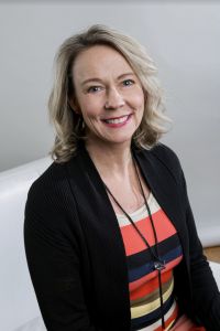 Kelly of Gold Coast Doulas poses in a white chair wearing a colorfully striped top and black blazer