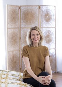 Kristin of Gold Coast Doulas sitting at the edge of a bed poses with her legs crossed and hands folded on top