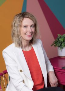 Kristin of Gold Coast Doulas sits in a pink chair at a pink desk with a plant in front of a colorful wall wearing a coral blouse and white blazer