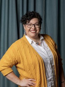 Kaytlyn of Gold Coast Doulas wearing a button up white shirt with a mustard yellow cardigan in front of a blue curtain