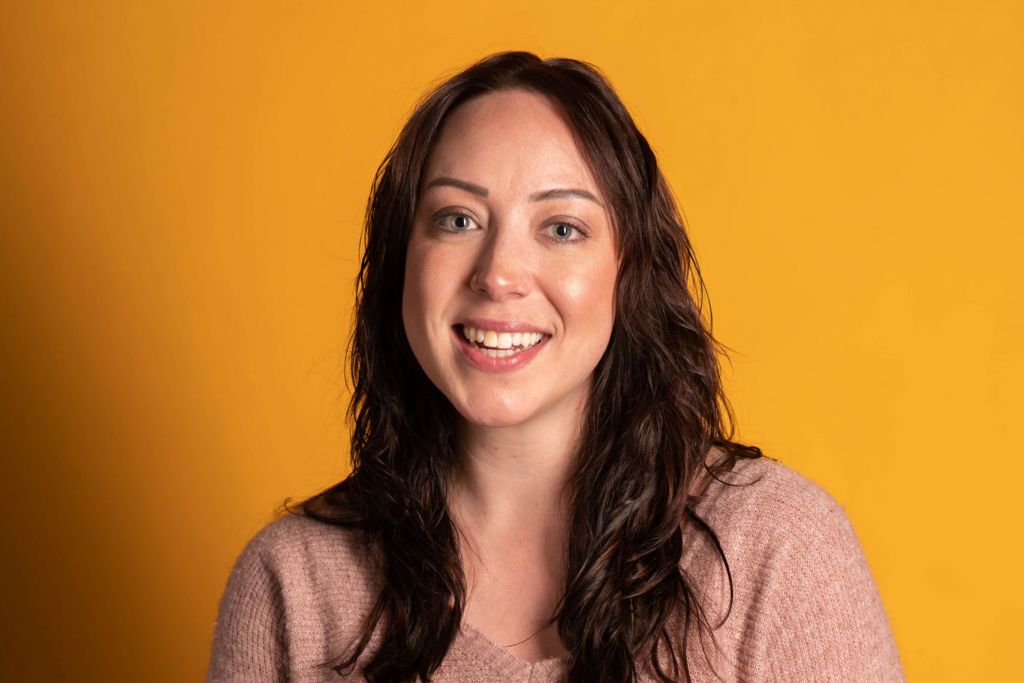 Deborah Kalsbeek poses in a pink sweater in front of an orange wall