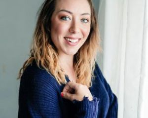 Woman wearing a navy blue sweater poses in front of a grey wall and white curtains