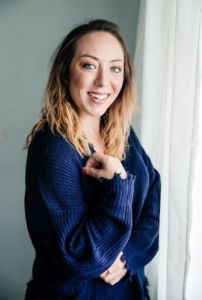 Woman wearing a navy blue sweater poses in front of a grey wall and white curtains