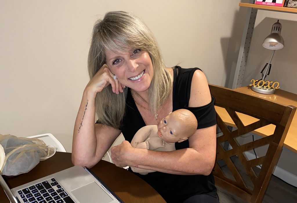 Gail holds a baby doll while sitting at a table with a laptop in front of her