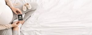Pregnant woman holding a sonogram image and baby shoes on a white bed