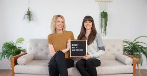 Kristin and Kay of Gold Coast Doulas holding a sign that says, "WE CAN'T WAIT TO HEAR FROM YOU!" on a grey couch with plants around them