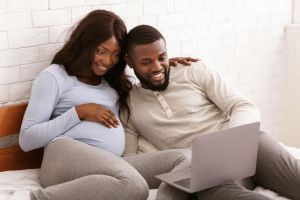 Pregnant couple snuggled together on a bed in front of a laptop