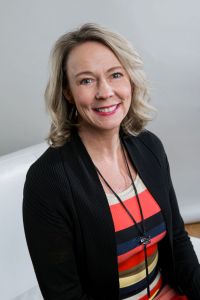 Kelly Emery poses on a white chair in front of a white wall wearing a striped top and black blazer