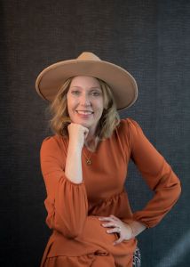 Kristin Birth Coaching poses in front of a black wall with her chin resting on her fist wearing an orange dress and hat