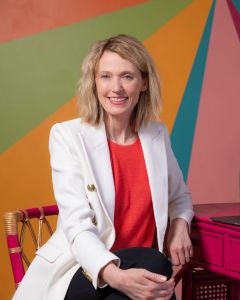 Kristin Revere poses at a pink desk wearing a red blouse and white blazer in front of a colorfully painted wall