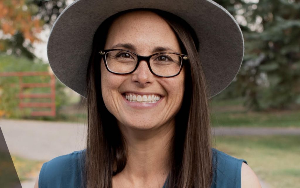 Sarah Thompson poses with a hat on, glasses, and blue tank top in front of trees