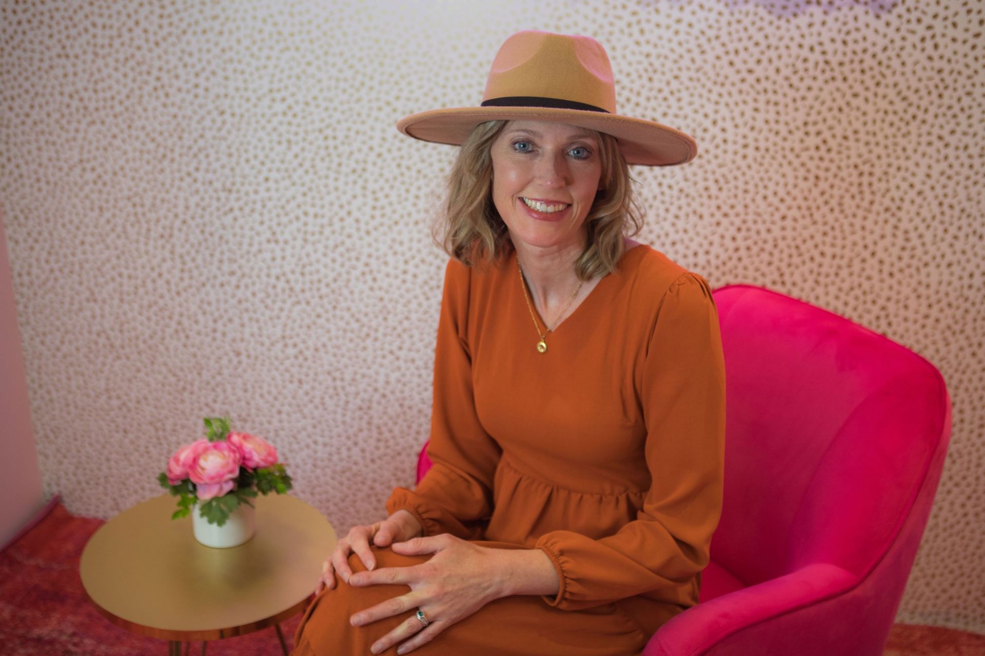 Birth Coaching with Kristin Revere poses in a pink chair wearing an orange dress and hat