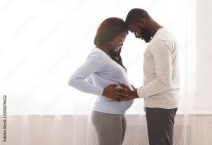 Couple facing one another smile and cradle the mother's pregnant belly in front of a bright and white window