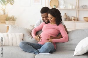 Pregnant woman wearing a pink sweater and jeans sits on a grey sofa with a man behind her cradling her pregnant belly and smiling together in a living room