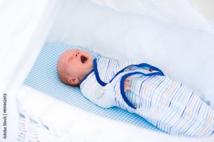 Yawning newborn in a blue striped Velcro swaddle rests in a blue and white bassinet