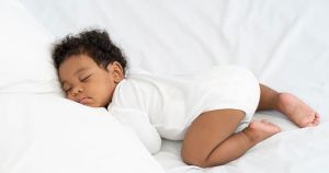Newborn baby sleeping on their tummy wearing a white long sleeve onesie on a soft white bed
