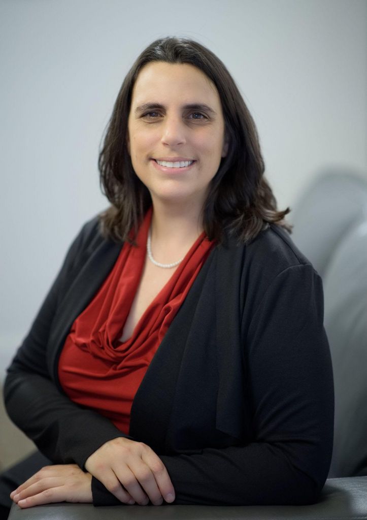 Cara wearing a red blouse, white pear necklace, and black blazer poses in a grey chair