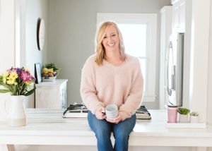 English Goldsborough wearing a fuzzy pink sweater sits on top of a counter wearing blue jeans and holding a mug in her hands