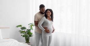 Smiling couple cradles the mother's pregnant belly in a bright bedroom with a green plant