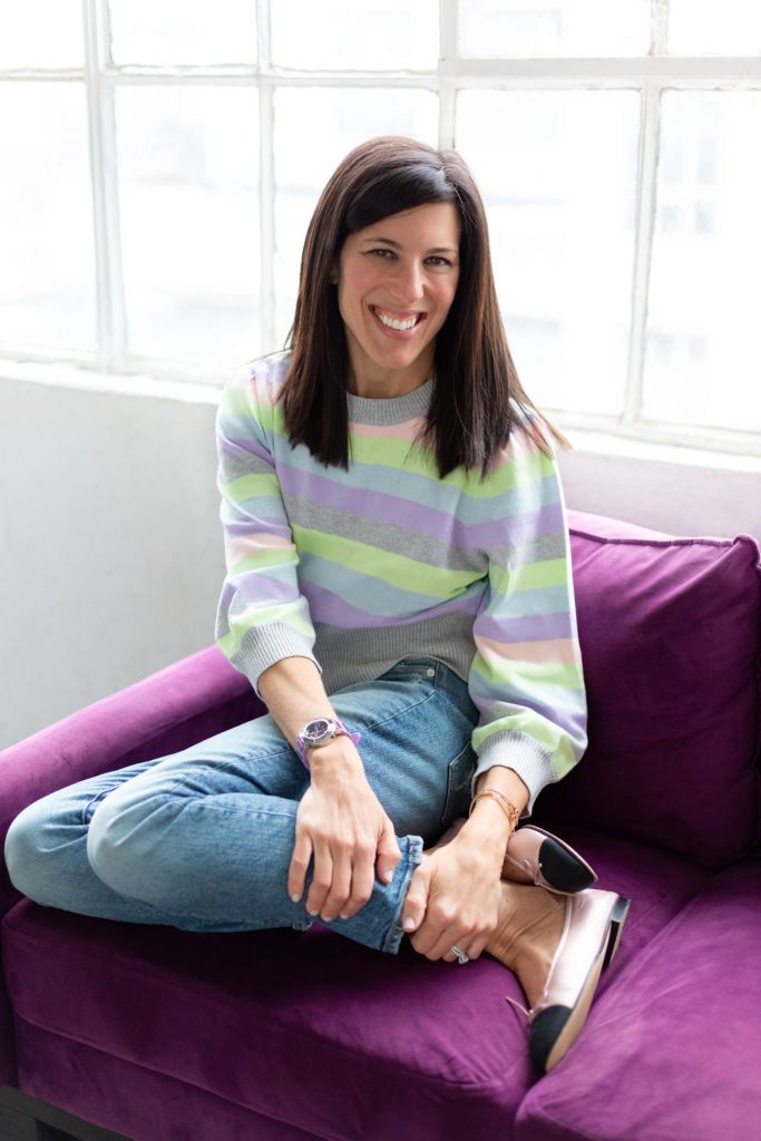 Jodi wearing a striped sweater, blue jeans, and ballet slippers sits on a purple couch in front of a window