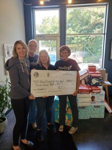Owner, Kristin Revere, of Gold Coast Doulas picking up a large check for the Nestlings Diaper Bank with three others surrounding her and boxes of diapers in the background