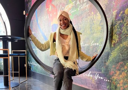 Mya from Gold Coast Doulas sits inside a floating circle in front of a Kitchen + Kocktale wall wearing a headband, scarf, and crossbody fanny pack.