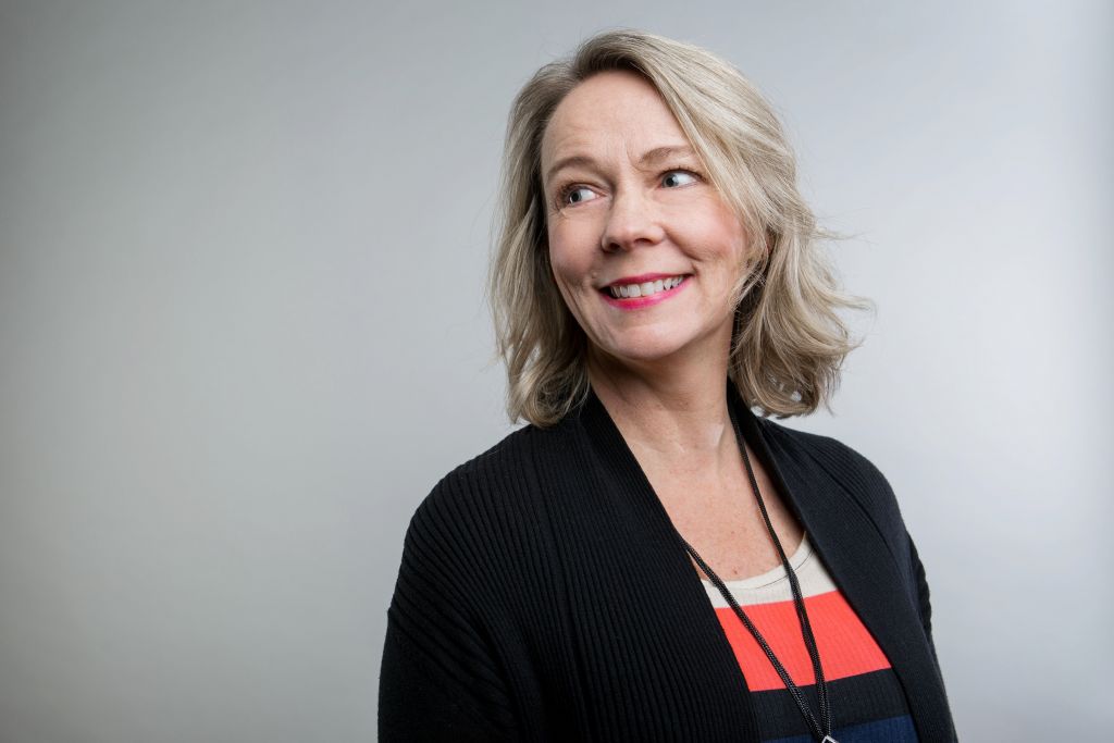 Professionally dressed woman with striped shirt and black cardigan smiling and looking over her shoulder in front of a grey wall