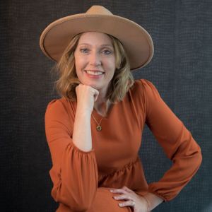 Kristin Revere wearing a burnt orange dress and a tan hat posing in front of a black alligator backdrop with her elbow on her knee and chin resting on her fist