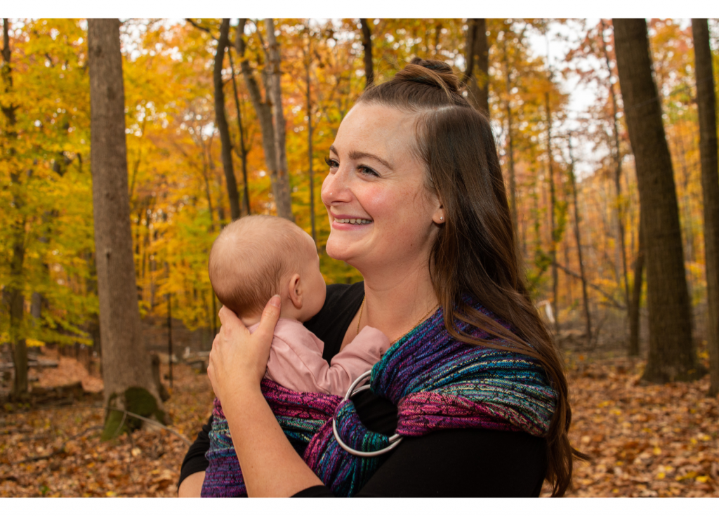 Lexi wearing black while wearing a baby in a colorful ring sling in an autumn forest