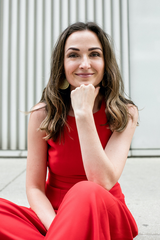 Magdalena Lasota-Morales wearing a red jumper, sitting on the ground, resting her head on her fist, with a grey wall behind her