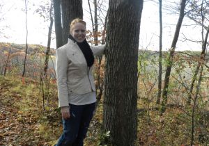 Woman standing by a tree in a wooded area with jeans and beige jacket