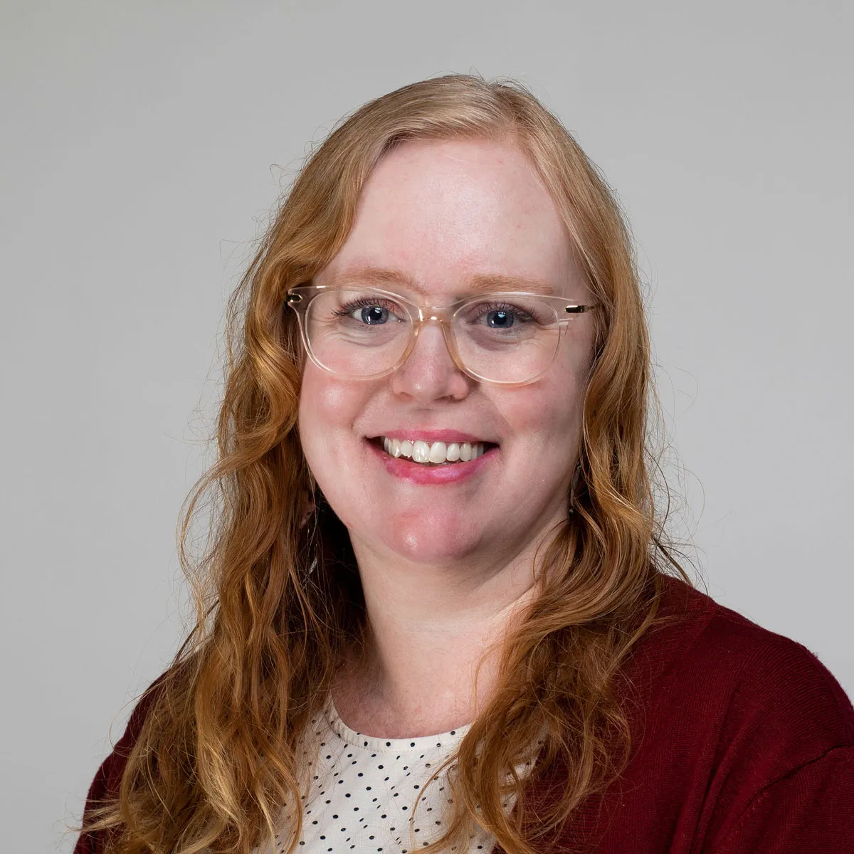 Sarah Short wearing a black and white polka dot blouse and red cardigan against a grey background