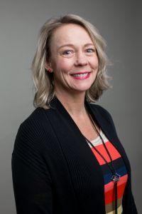 Professionally dressed woman wearing a striped shirt, black cardigan, and long necklace poses for a headshot in front of a grey background