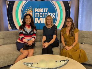 Three women professionally clothed sitting on a rounded couch in front of a Fox17 Morning MI screen