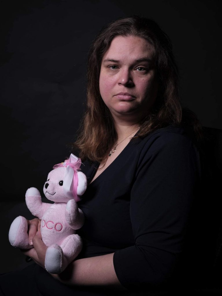 Michelle wearing a black top while holding a pink teddy bear against a black wall