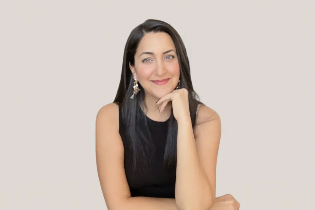 Woman poses in a black sleeveless dress with her hand resting on her chin against a grey/purple background