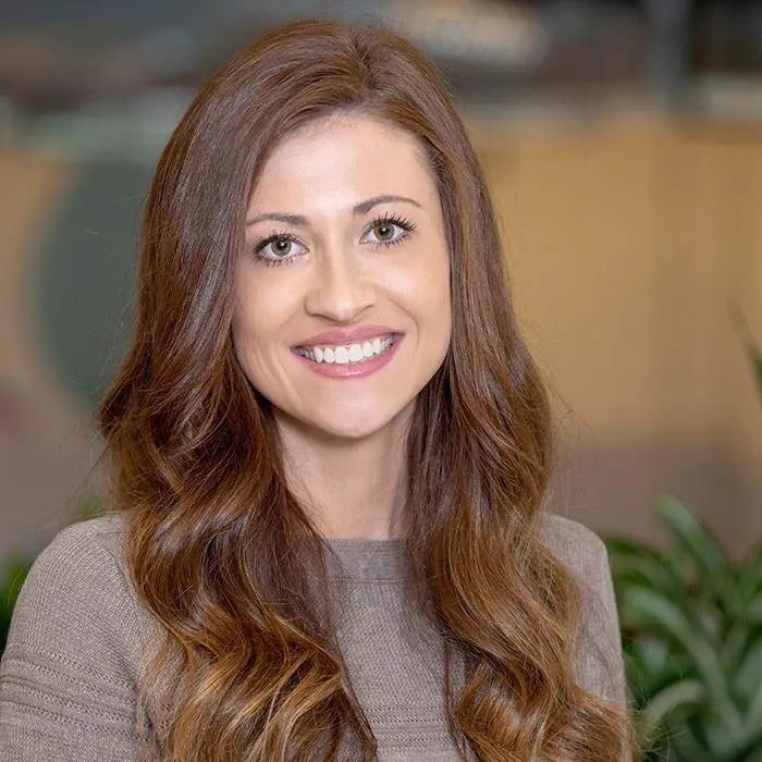 Headshot of a woman wearing a brown sweater with office and plants in the background