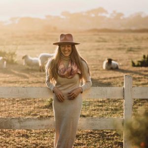 Dr. Laura wearing a long beige sweater dress with plaid scarf and hat leaning against a white fence with sheep in the background