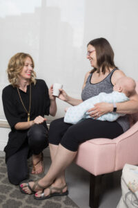 Mom holds her baby in a chair while Kristin from Gold Coast Doulas hands her a mug
